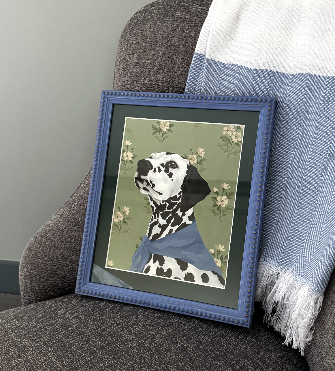 Framed artwork of a dalmatian dog on a gray armchair with a blue blanket.