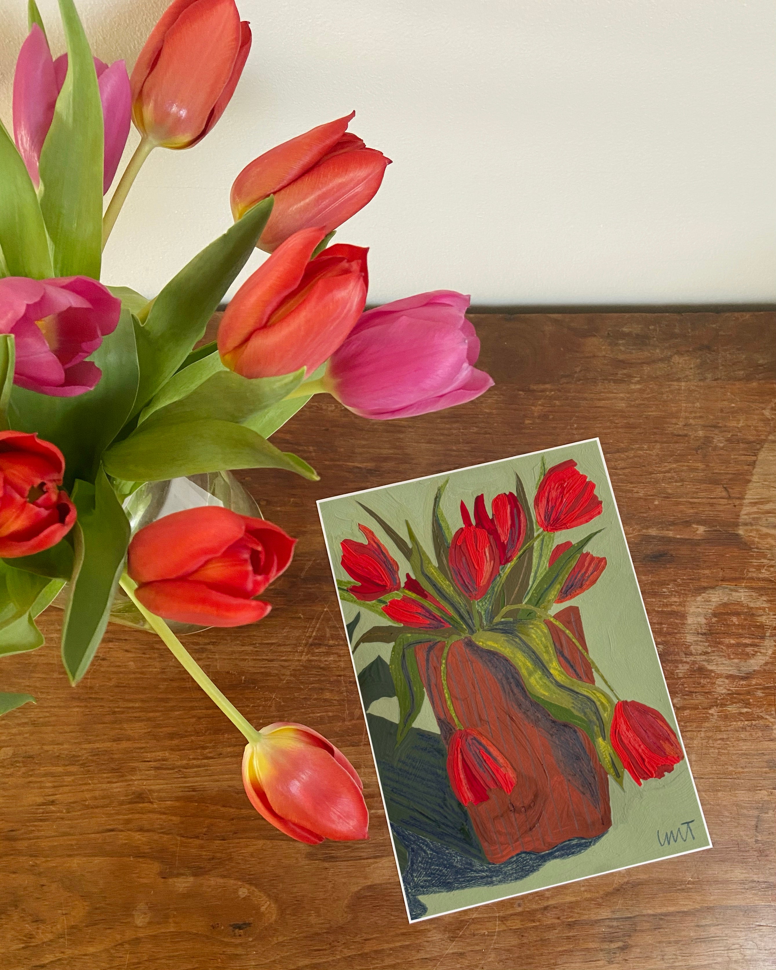 Bouquet of red and pink tulips on a wooden surface with a matching postcard.